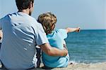 Vater und Sohn sitzen zusammen am Strand, junge zeigt auf See