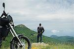 Man standing on rock looking at mountain view, motorcycle in foreground, rear view