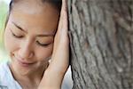 Woman resting head against tree trunk with eyes closed