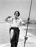 1920s - 1930s SMILING WOMAN STANDING IN OCEAN SURF WEARING RUBBER WADERS HOLDING FISH AND FLY FISHING ROD