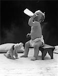 1930s BABY SEATED ON STOOL DRINKING MILK FROM BOTTLE WITH STUFFED PUPPY LOOKING UP AT HIM