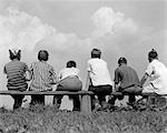 1960s BACK VIEW OF SIX BOY BASEBALL PLAYERS SITTING ON BENCH