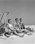1960s FAMILY PORTRAIT MOTHER FATHER DAUGHTER AND TWO SONS SITTING ON BEACH UNDER UMBRELLA
