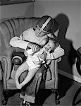 1950s - 1960s BOY SITTING BY FOOTBALL IN EASY CHAIR WEARING HELMET TRYING TO HOLD AND FEED BOTTLE TO CRYING BABY