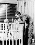1930s - 1940s - 11 MONTH OLD BABY STANDING IN CRIB IN FRONT OF WINDOW WITH VENETIAN BLINDS FATHER HANDING BABY A TOY DOG STUFFED ANIMAL NURSERY