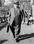 1960s OVERWEIGHT BUSINESSMAN WITH BRIEFCASE SMOKING CIGAR WALKING ON CITY SIDEWALK