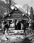 ANNÉES 1920 - ANNÉES 1930 COUPLE DEVANT LA CABANE EN RONDINS PERMANENT PAR DES CHEVAUX AVEC SELLE PORT CANADA JODHPURS ASSINIBOINE