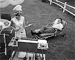 1950s - 1960s WOMAN IN CHEF'S HAT AND APRON GRILLING HOT DOGS ON GRILL MAN IN CHAISE LONGUE LAUGHING EATING A FRANKFURTER OUTSIDE