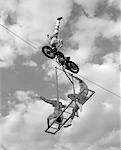 1950s - 1960s HIGH-WIRE ACT WITH MAN STANDING ON HIS HEAD ON MOTORCYCLE WITH MAN AND WOMAN ON BALANCED LADDER SUSPENDED BELOW