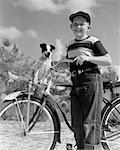 ANNÉES 1940 - ANNÉES 1950 BOY SUR VÉLO AVEC PUPPY IN BASKET REGARDANT LA CAMÉRA