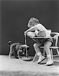 1940s TODDLER SITTING IN CHAIR POURING MILK FROM BOTTLE INTO BOWL FOR PUPPY DOG