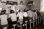 1950s REAR VIEW OF GROUP OF TEENAGE BOYS & GIRLS SITTING TOGETHER AT A SODA FOUNTAIN MALT SHOP COUNTER SNACK FOOD