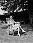 1940s WOMAN SITTING IN WOODEN LAWN CHAIR WITH WHEELS ON LAWN IN FRONT OF HOUSE LOOKING OFF INTO DISTANCE