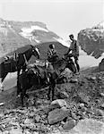 ZWANZIGER - DREIßIGER JAHRE PAAR MANN FRAU TRAGEN RIDING GEAR REITHOSE STIEFEL SPOREN SITZEND STANDING ON GROßER FELSEN VON ZWEI PFERDEN