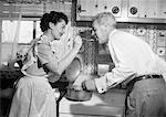 1950s HOUSEWIFE IN KITCHEN HAVING HUSBAND TASTE FOOD ON STOVE