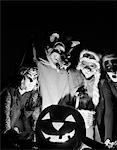 1960s GROUP OF FOUR CHILDREN IN HALLOWEEN COSTUMES GATHERED AROUND JACK-O'-LANTERN INDOOR