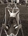 1950s RELAXED SMILING BOY LYING IN SUBURBAN BACKYARD HAMMOCK