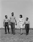 1960s AFRICAN-AMERICAN FAMILY HOLDING HANDS WALKING IN GRASSY FIELD