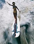 ANNÉES 1960 FEMME EN MAILLOT DE BAIN DEBOUT ÉQUILIBRAGE SURFANT SUR UNE PLANCHE DE SURF