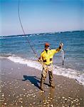 1970s MAN RED CAP HAT YELLOW SHIRT WADERS ON BEACH SURF FISHING HOLDING UP CATCH OF THE DAY