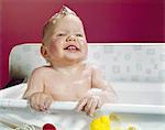 1960s SMILING BABY HOLDING ON TO SIDE OF INFANT BATH