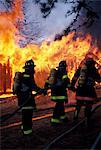 FIREMEN WITH BREATHING EQUIPMENT FULL GEAR ADVANCING WITH HOSE ON FLAMES IN BACKGROUND