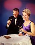 DES ANNÉES 1960 - ANNÉES 1970 COUPLE HOMME ET FEMME EN FORMELLE SMOKING ET ROBE DU SOIR SOURIANT GRILLER AVEC VERRES DE CHAMPAGNE