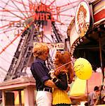 1970s YOUNG MAN WOMAN HUGGING BY CARNIVAL RIDES HOLDING BALLOON WONDER WHEEL OF CONEY ISLAND