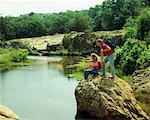 1980s YOUNG COUPLE HIKING ON ROCK BY POND