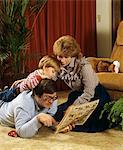 1980s FAMILY ON LIVING ROOM FLOOR FATHER READING NEWSPAPER COMICS TO BOY AND MOTHER