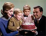 1960s MOTHER AND FATHER HOLDING BIRTHDAY CAKE AND SONS BLOWING OUT CANDLES