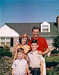 1960s PORTRAIT SMILING FAMILY FATHER MOTHER TWO SONS STANDING TOGETHER IN FRONT OF SUBURBAN HOUSE
