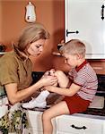 1970s MOTHER GIVING FIRST-AID PUTTING BAND-AID ON KNEE OF SON IN KITCHEN