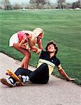 1970s LAUGHING TEEN GIRL HELPING BOY UP AFTER HE FELL OFF SKATEBOARD