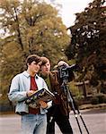 1960s TWO TEEN BOYS ONE OPERATING MOVIE CAMERA THE OTHER HOLDING SCENE MARKER OUTDOORS