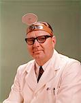 1950s - 1960s SMILING MAN MEDICAL DOCTOR WHITE LAB COAT BLACK FRAME EYEGLASSES WEARING DIAGNOSTIC HEAD MIRROR ON FOREHEAD