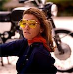 1970s SEXY YOUNG WOMAN WITH LONG AUBURN HAIR WEARING YELLOW SUNGLASSES SITTING BY A MOTORCYCLE