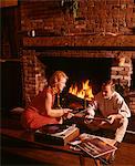 1960s - 1970s COUPLE MAN WOMAN BY LIVING ROOM FIREPLACE PLAYING VINYL RECORD ALBUM MUSIC ON HIGH FIDELITY STEREO TURNTABLE