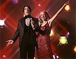 1970s DUO MAN WOMAN SINGING WOMAN RED DRESS GOWN MAN TUXEDO HOLDING MICROPHONE STAGE LIGHTS BACKGROUND