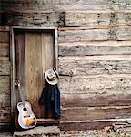 1960s - 1970s GUITAR, HAT AND JACKET HANGING BY WEATHERED LOG CABIN DOOR