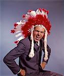 DES ANNÉES 1960 EN COLÈRE HOMME HOMME D'AFFAIRES DANS LA COIFFE DE GUERRE BONNET PLUME AMÉRINDIENNE ET COSTUME AVEC LES MAINS SUR LES HANCHES