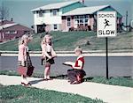 1950s TWO TWIN BLOND GIRLS ONE BLOND BOY ON SIDEWALK BY SCHOOL SLOW SIGN IN NEIGHBORHOOD OF SUBURBAN SPLIT LEVEL HOUSES