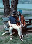 1970s YOUNG COUPLE SITTING ON PARK BENCH WEARING GAS MASKS