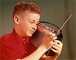 1960s SMILING YOUNG RED HAIR PRE-TEEN BOY LISTENING TO PORTABLE TRANSISTOR RADIO HELD UP TO EAR