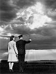 ANNÉES 1940 COUPLE HOMME FEMME POINTANT AU COUCHER DU SOLEIL, MAIN DANS LA MAIN DE L'HOMME PORTANT UN UNIFORME MILITAIRE