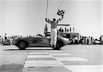 ANNÉES 1960 HOMME SAUTANT AGITANT CHECKERED FLAG AS REMPORTÉ LA VOITURE DE SPORT, FRANCHISSANT LA LIGNE D'ARRIVÉE