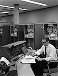 HOMME DES ANNÉES 1960 DANS LA CHEMISE & TIE & VERRES AU PUPITRE DE COMMANDE POUR SYSTÈME DE TRAITEMENT DE DONNÉES D'IBM