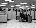 1960s MAN TECHNICIAN PROGRAMMING MAINFRAME COMPUTER AT CONTROL ROOM CONSOLE SURROUNDED BY MANY TAPE DRIVE DATA STORAGE MACHINES