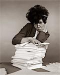 1960s - 1970s WOMAN SECRETARY PENCIL STUCK IN TEASED HAIR OVERWORKED EXPRESSION LEANING PILE PAPERWORK