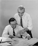 1960s 2 BUSINESSMEN IN SHIRT SLEEVES LOOKING OVER PAPERWORK